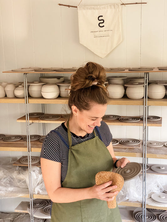 Potter Sara Scheuermann in the Steaped Slow Ceramics studio with shelves of handmade pottery in progress and a banner with the Steaped Slow Ceramics logo.