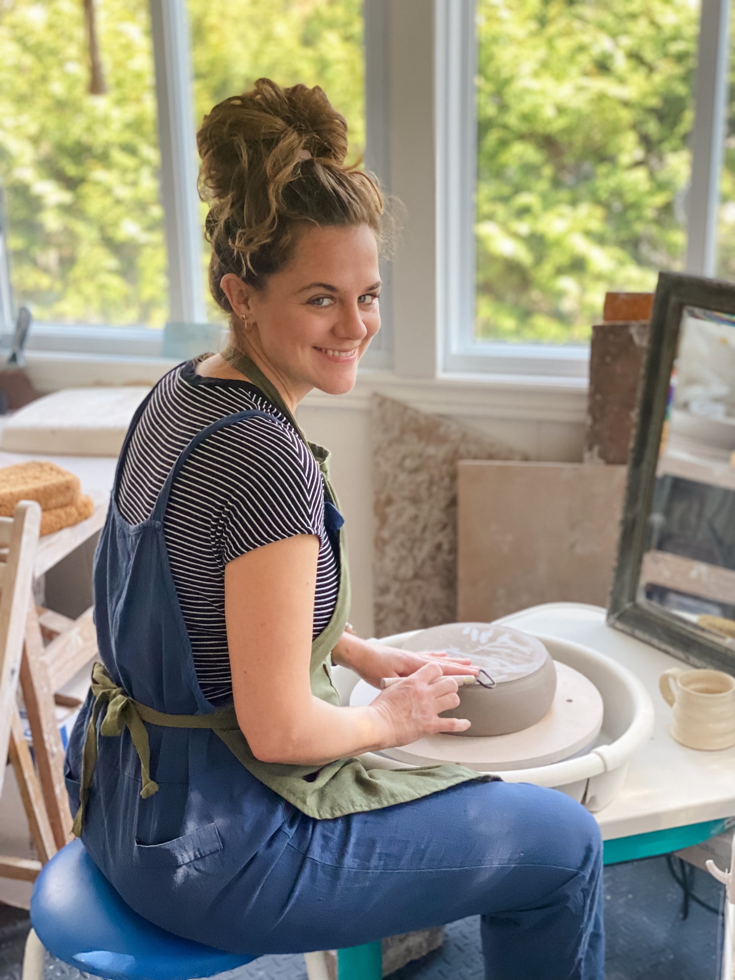 Potter, Sara Scheuermann sitting behind the potter's wheel in her bright studio