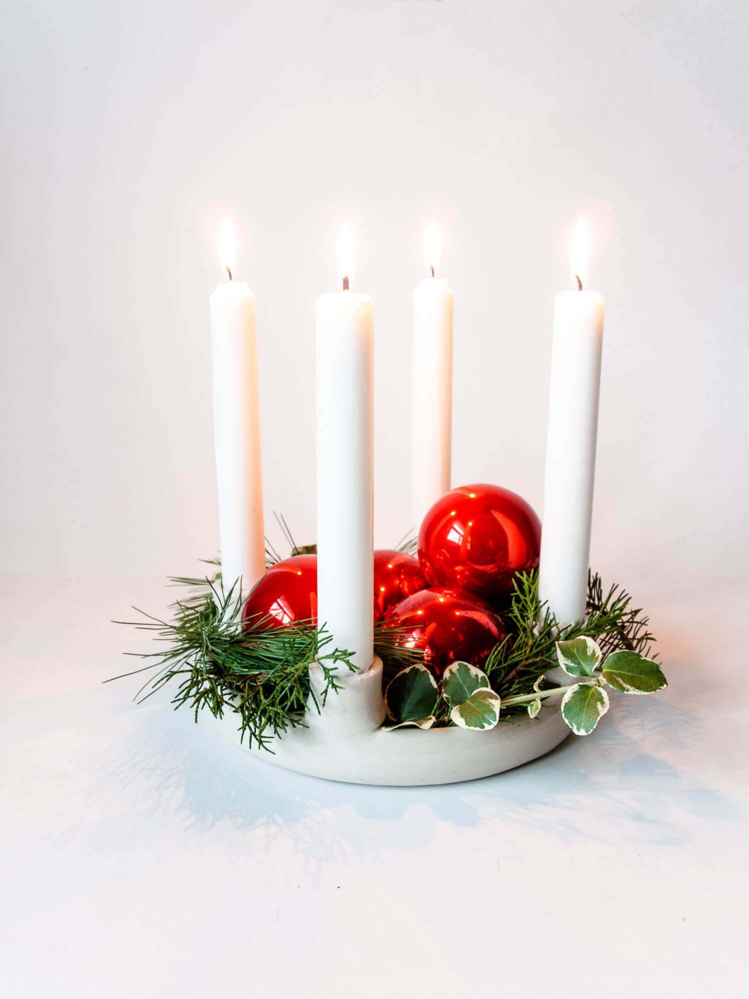 White pottery advent wreath candle holder, with four lit tapered candles, living green cedar, pine and holly foliage and red glass Christmas bulbs for celebrating advent tradition.