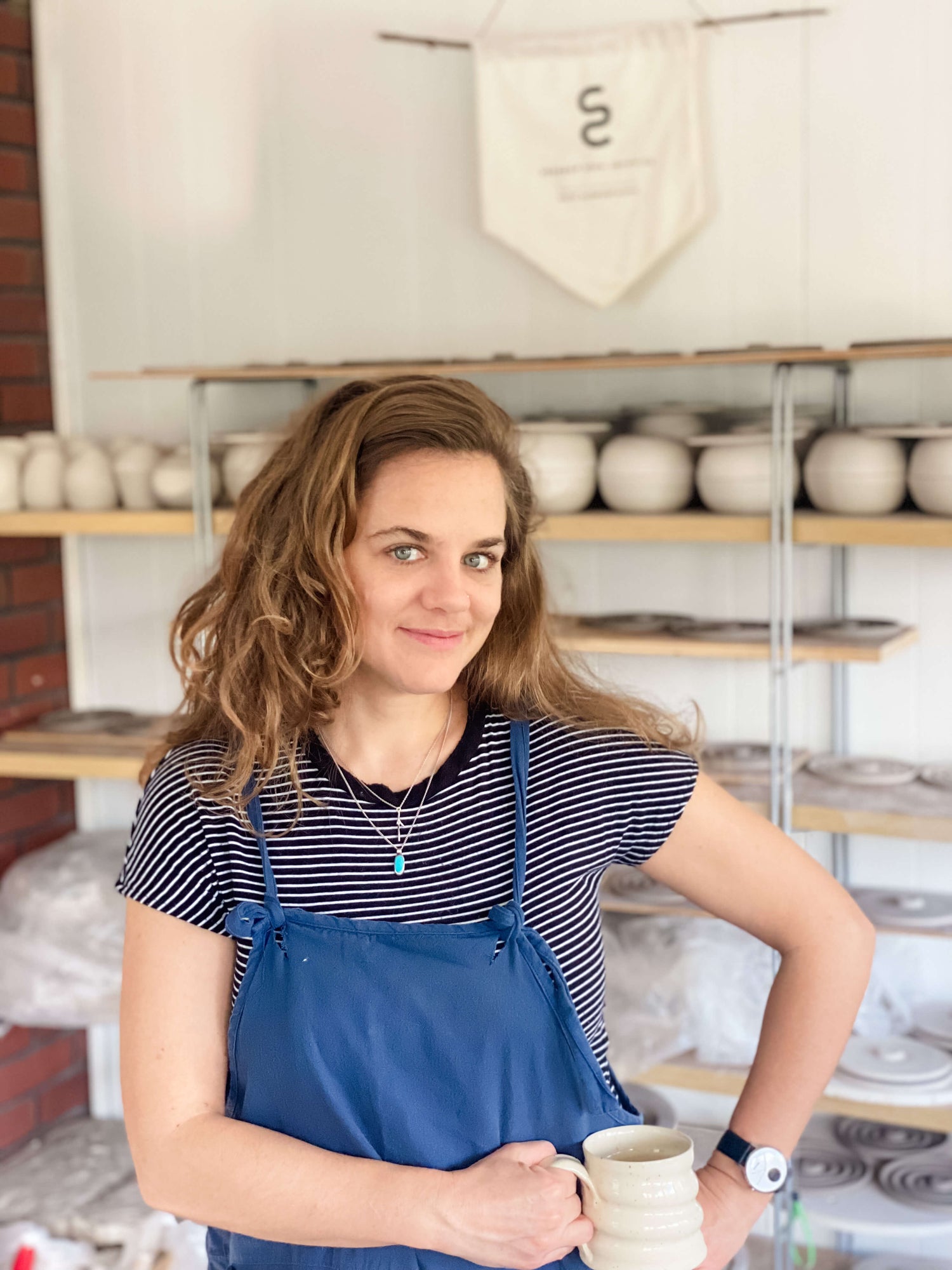 Artist and potter Sara Scheuermann in studio standing in front of a shelf full of pottery, holding a handmade mug and smiling at the camera 