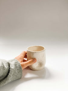 Hand holding wheel thrown tumbler in cream clay with speckles with two dimples pinched into the sides