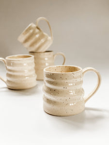 wheel thrown mug with pronounced spiral throwing marks in cream and brown speckled clay with 3 in mugs in the background 
