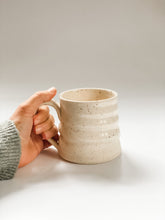 Load image into Gallery viewer, Hand holding wheel thrown mug with pronounced spiral throwing marks in cream and brown speckled clay  