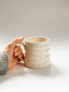 Hand holding wheel thrown mug with pronounced spiral throwing marks in cream and brown speckled clay  