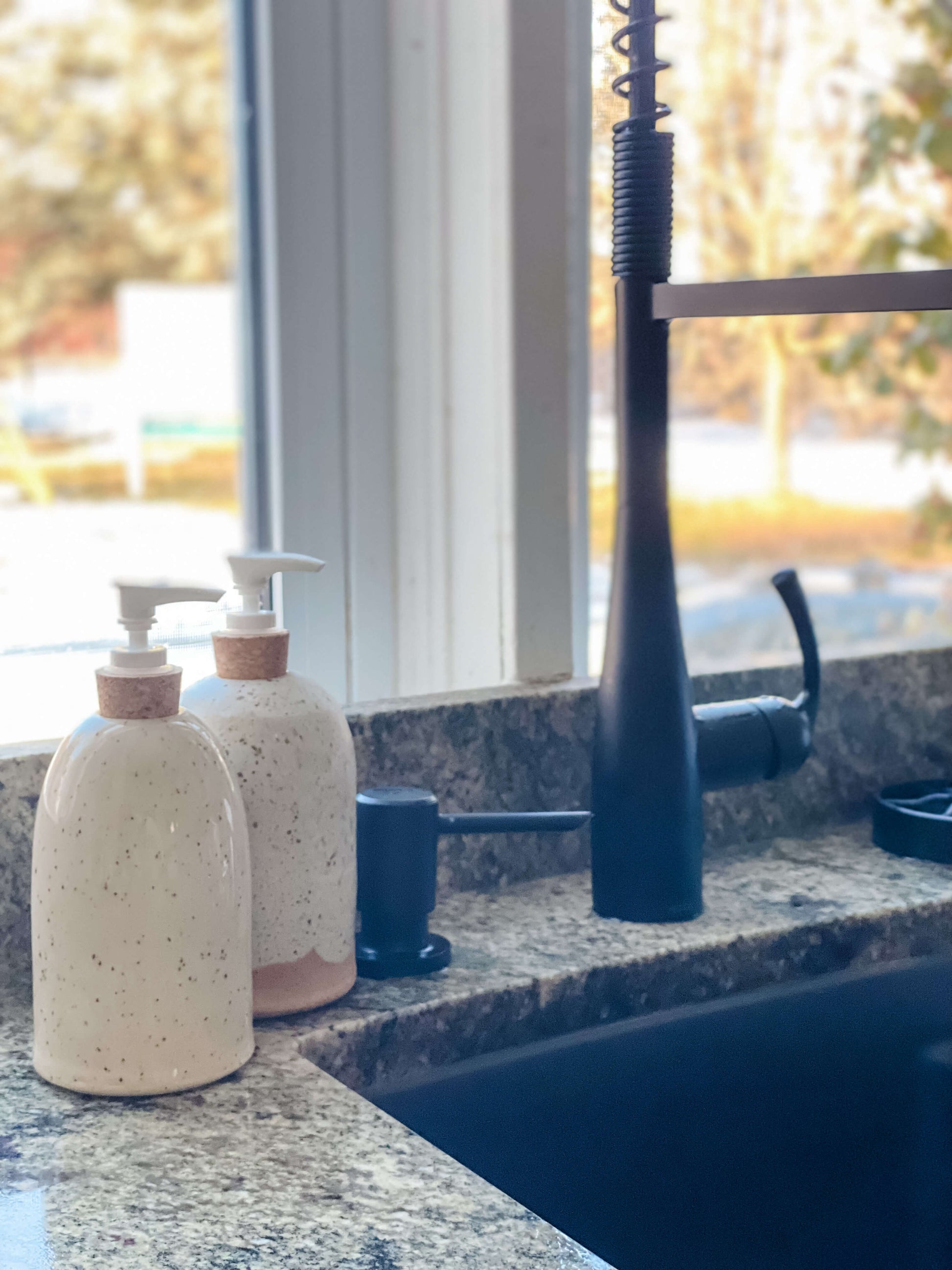 Two white and cream stoneware pottery soap pumps beside a kitchen sink with a dark grey granite counter top