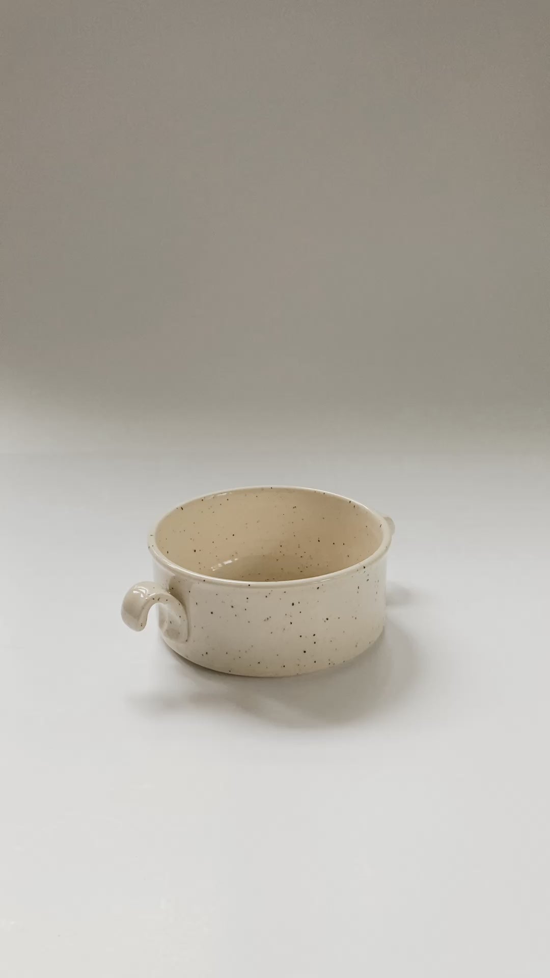 Hands picking up and showing off a wheel thrown cylindrical soup bowls with two hooked handles in a cream and brown speckled clay